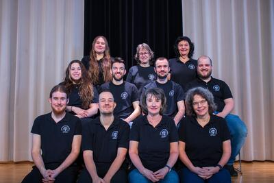 Gruppenfoto des Theaterverein Mönchaltorf in Mönchaltorf (8617, Kanton Zürich). Die Mitglieder des Vereins stehen und sitzen gemeinsam auf einer Bühne vor einem Bühnenvorhang und tragen einheitliche schwarze Vereinskleidung mit Vereinslogo. Das Bild zeigt die aktive Theatergruppe mit Mitgliedern verschiedener Altersgruppen bei einer offiziellen Aufnahme.