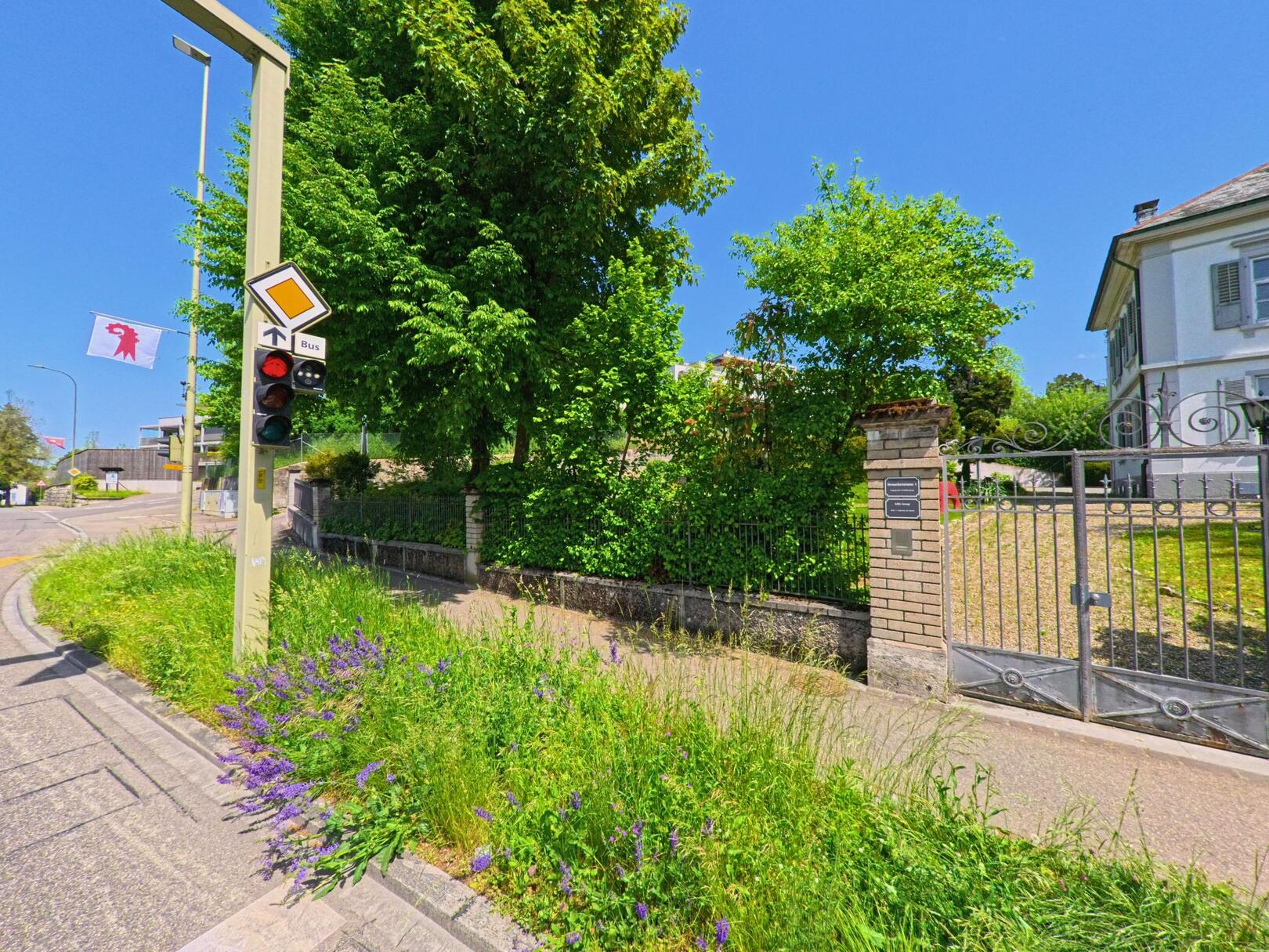 In Therwil (4106, Kanton Basel-Landschaft) steht eine Bushaltestellenampel an einer Kreuzung neben einem alten Wohnhaus mit schmiedeeisernem Tor.