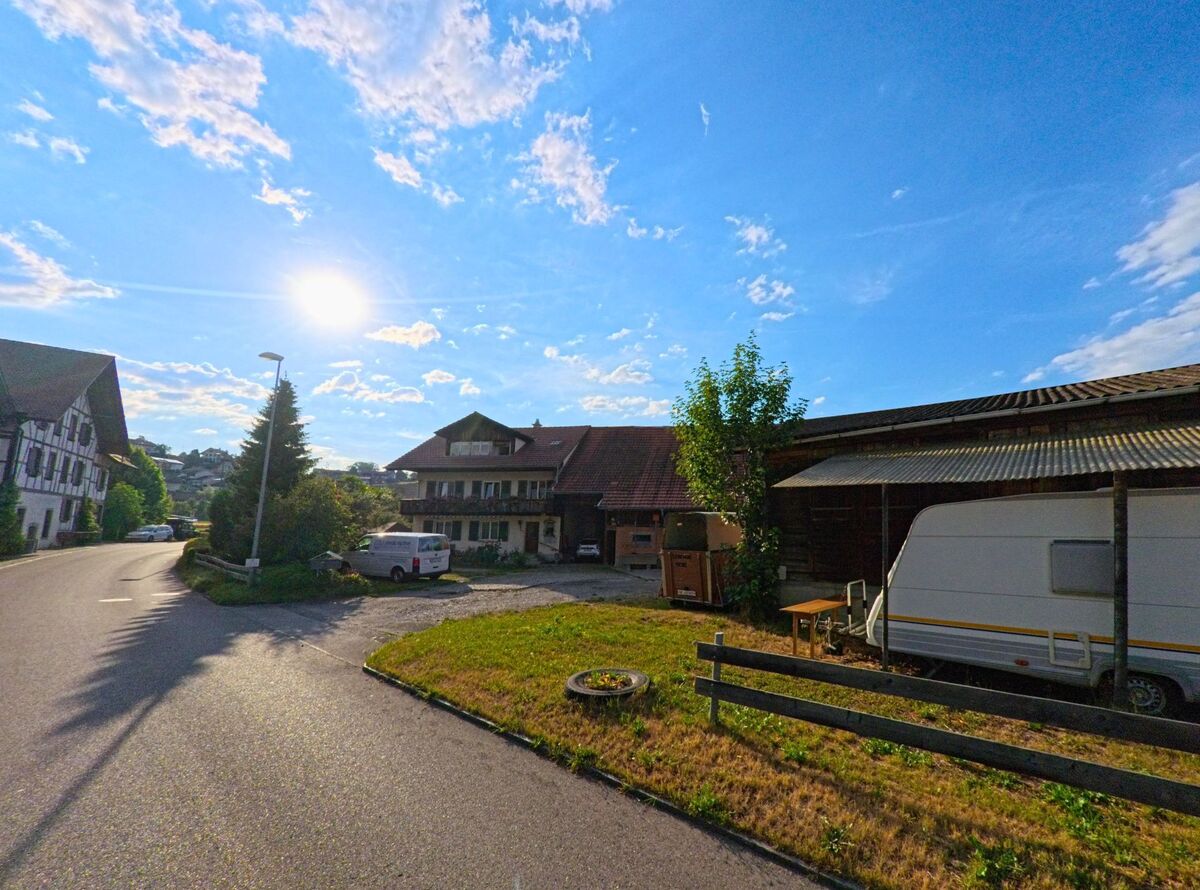 In Thierachern (3634, Kanton Bern) stehen ein Bauernhaus mit Holzfassade und ein Wirtschaftsgebäude mit abgestelltem Wohnwagen an einer ruhigen Strasse bei tiefstehender Sonne.