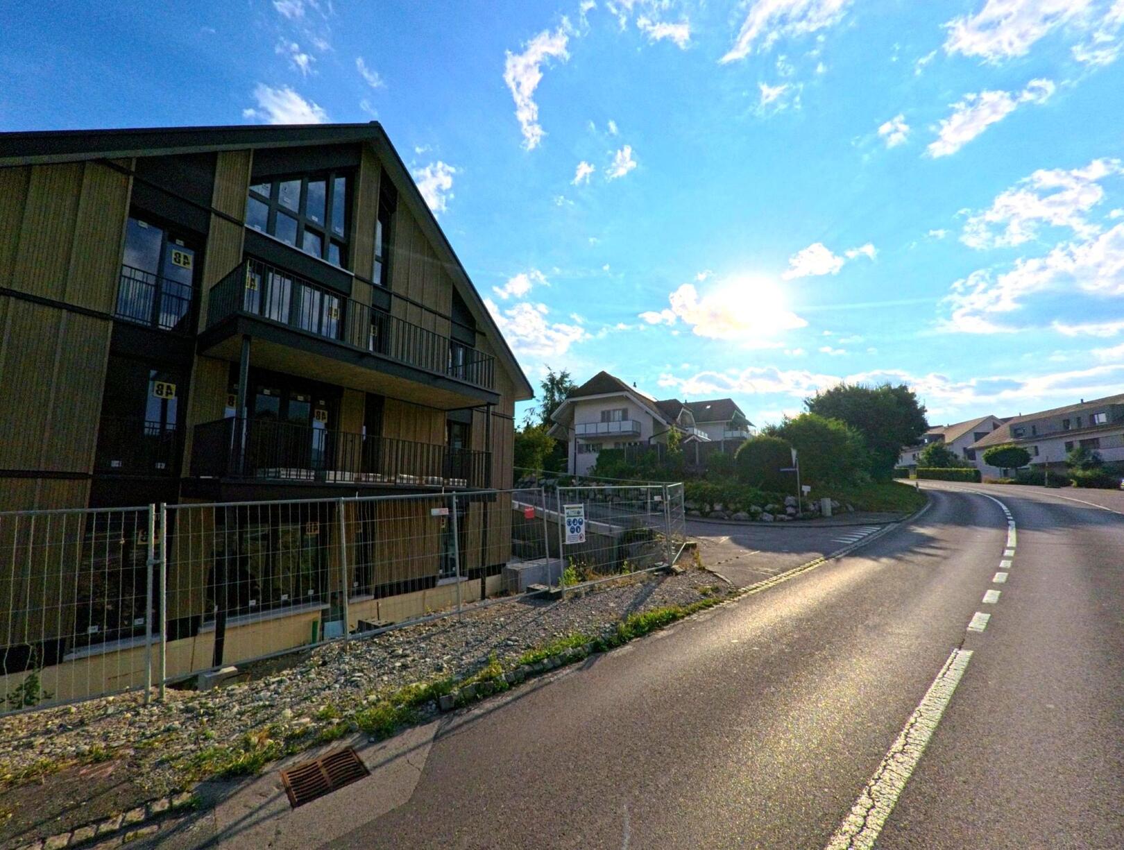 In Thierachern (PLZ 3634, Kanton Bern) verläuft eine Strasse durch ein Wohnquartier mit modernen Neubauten, während die tiefstehende Sonne für eine kontrastreiche Abendstimmung sorgt.