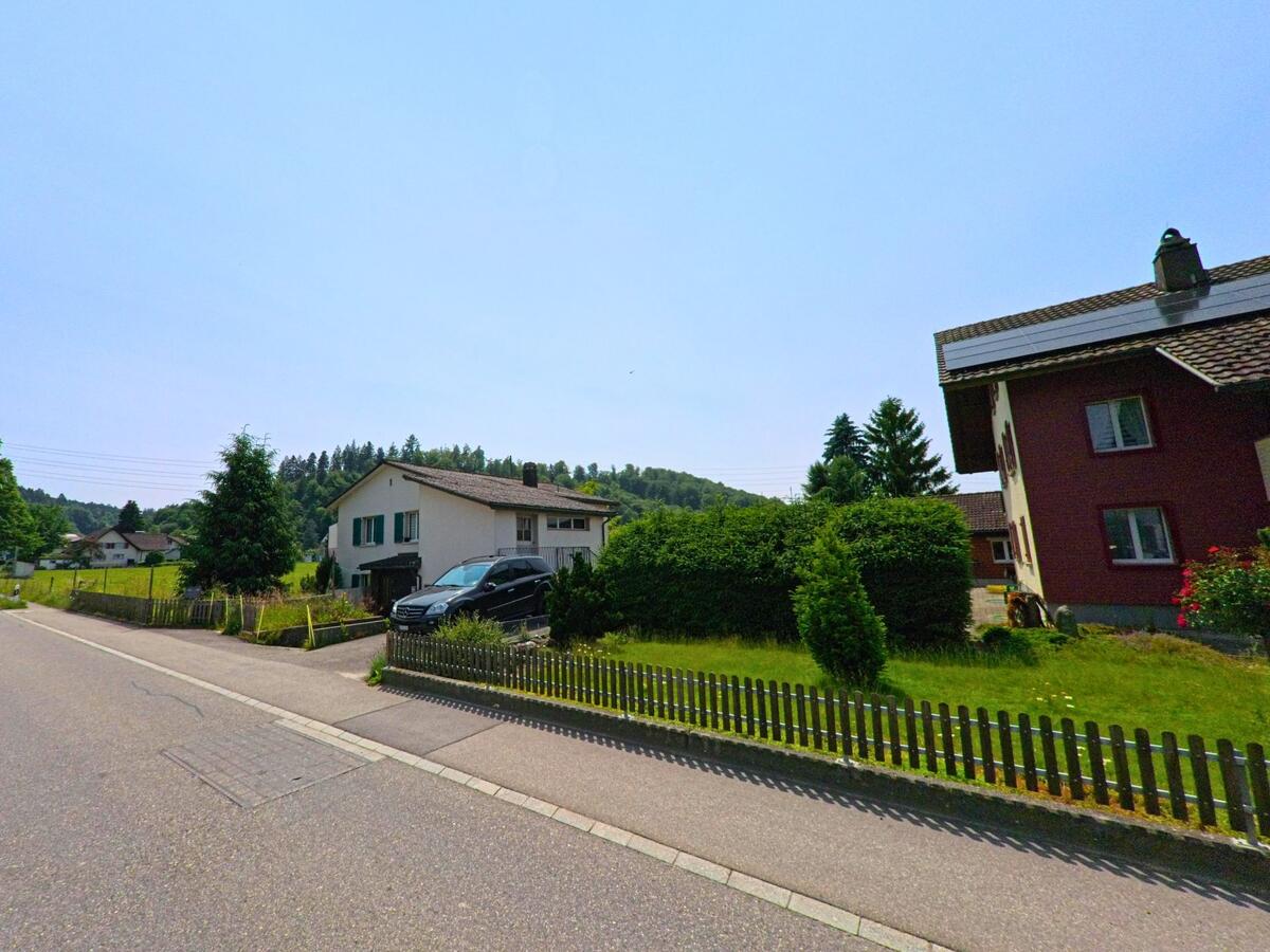In Thörigen (3367, Kanton Bern) reihen sich Einfamilienhäuser mit Gärten und Zäunen entlang einer wenig befahrenen Quartierstrasse am Waldrand.