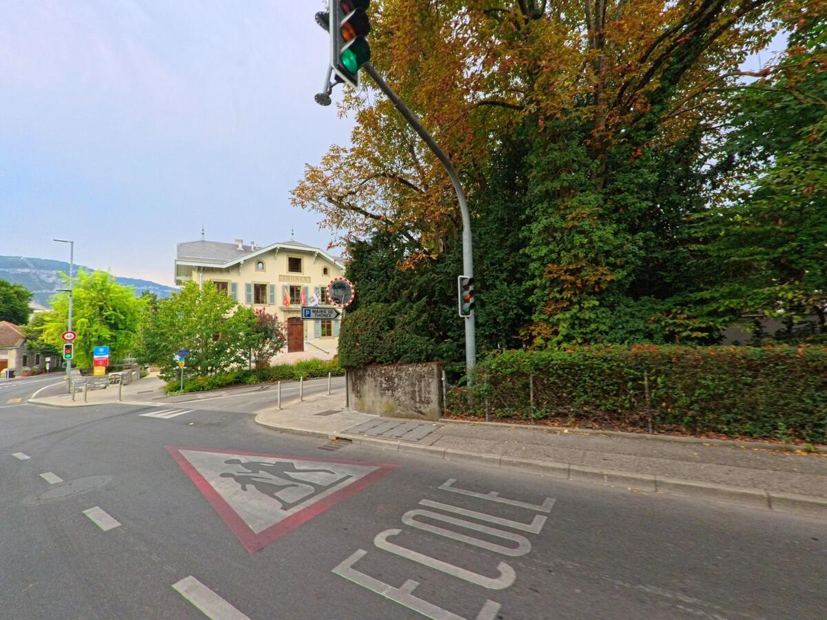 Vor dem Rathaus von Thonex (1226, Kanton Genf) fuehrt eine Strasse mit Schulwegmarkierung und Ampel an grossen Baeumen vorbei.