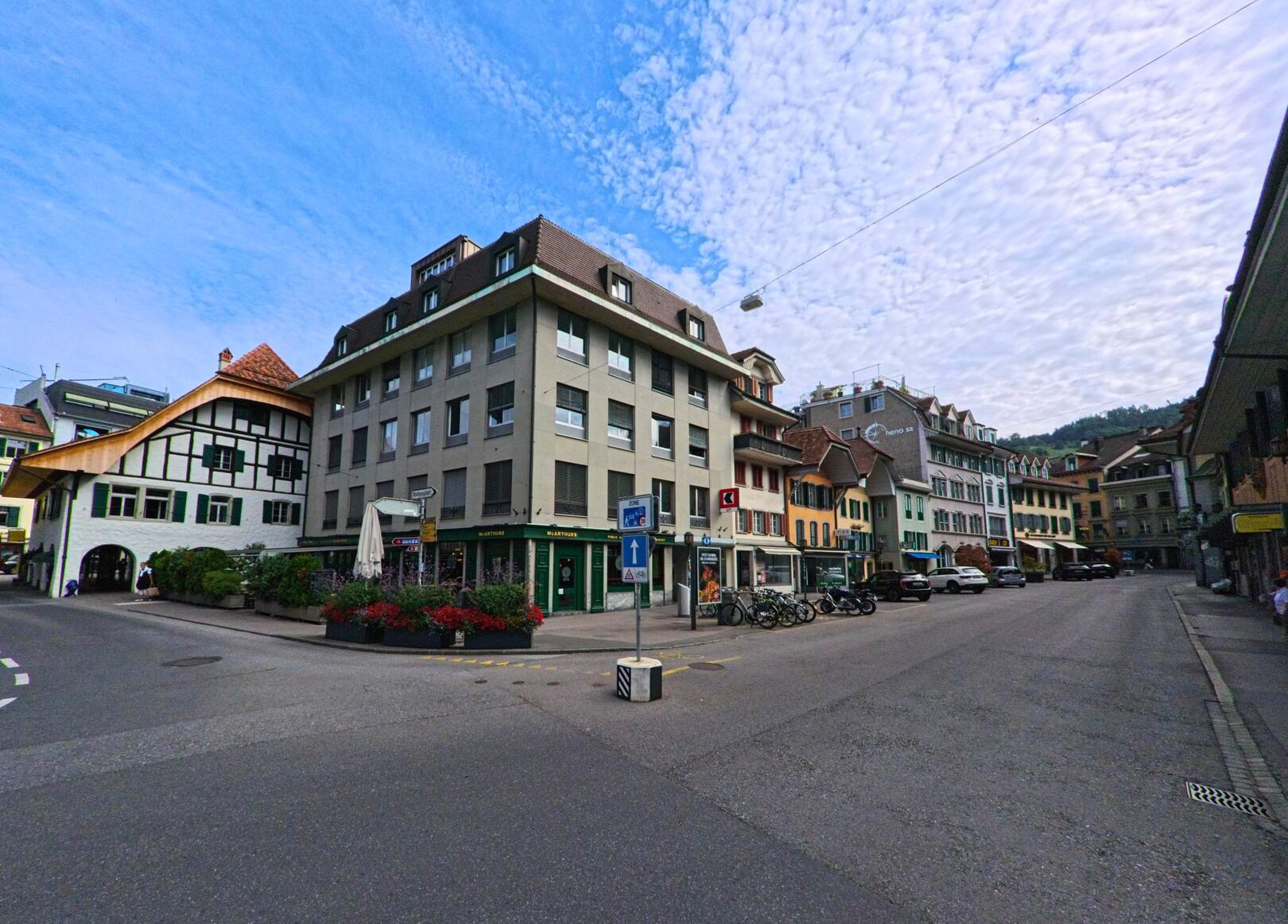 In Thun (3600, Kanton Bern) treffen moderne Wohnhäuser und traditionelle Fachwerkbauten an einer ruhigen Kreuzung mit Blumenbeeten und Fahrradständern aufeinander.