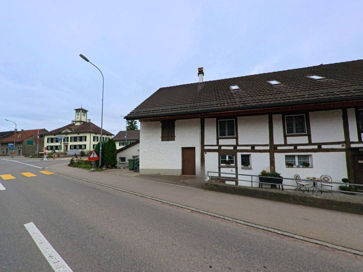 Entlang einer Hauptstrasse praegt ein traditionelles Fachwerkhaus das Ortsbild von Thundorf (8512, Kanton Thurgau), waehrend im Hintergrund ein markantes Gebaeude mit Uhrturm sichtbar ist.