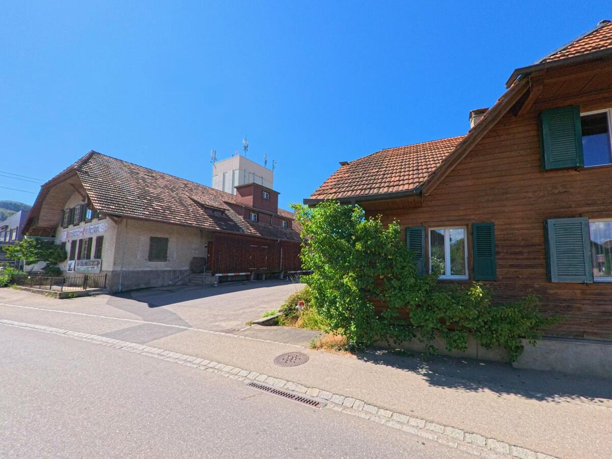 In Thurnen (3127, Kanton Bern) stehen entlang der Hauptstrasse traditionelle Wohnhäuser aus Holz, während im Hintergrund ein markanter Siloturm mit Mobilfunkantennen sichtbar ist.