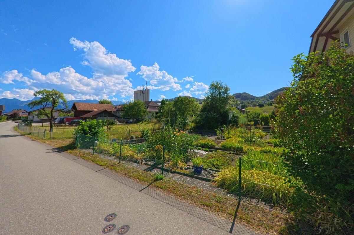 In Thurnen (3127, Kanton Bern) verläuft eine ruhige Quartierstrasse entlang von Hausgärten mit Blick auf den markanten Siloturm und die bewaldeten Hügel im Hintergrund.