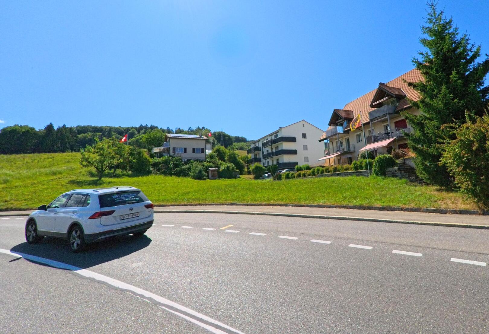 In Thurnen (3128, Kanton Bern) verläuft eine Hauptstrasse am sonnigen Ortsrand entlang, flankiert von Mehrfamilienhäusern mit Balkonen und gepflegten Grünflächen.