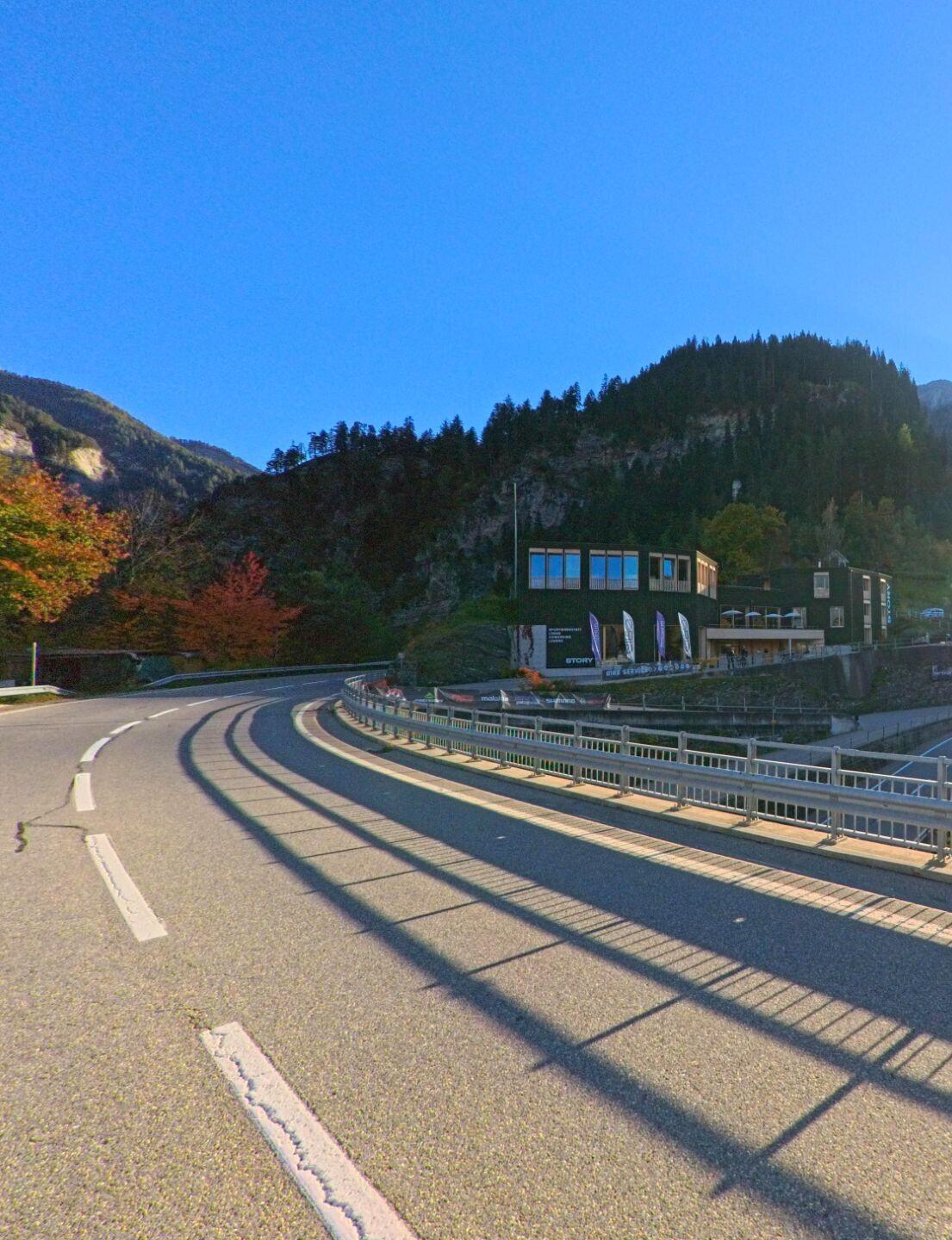 Entlang einer geschwungenen Brücke verläuft in Thusis (7430, Kanton Graubünden) eine Strasse mit Blick auf ein modernes Gebäude vor bewaldeten Hängen und herbstlicher Vegetation.