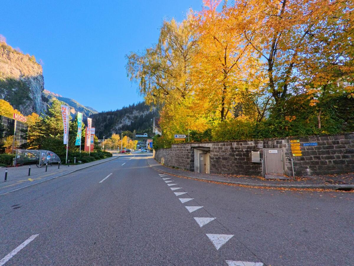 Entlang einer breiten Hauptstrasse führt der Blick in Thusis (7430, Kanton Graubünden) an herbstlich gefärbten Bäumen, Wegweisern und Geschäftsgebäuden vorbei in Richtung Ortszentrum.