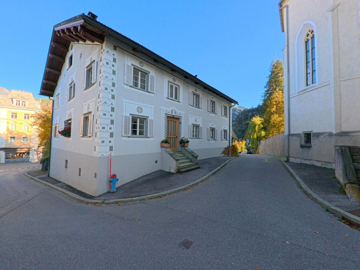 An einer schmalen Gasse steht in Thusis (7430, Kanton Graubünden) ein verziertes Wohnhaus mit Fensterläden, eingerahmt von historischer Bebauung und herbstlichen Bäumen.