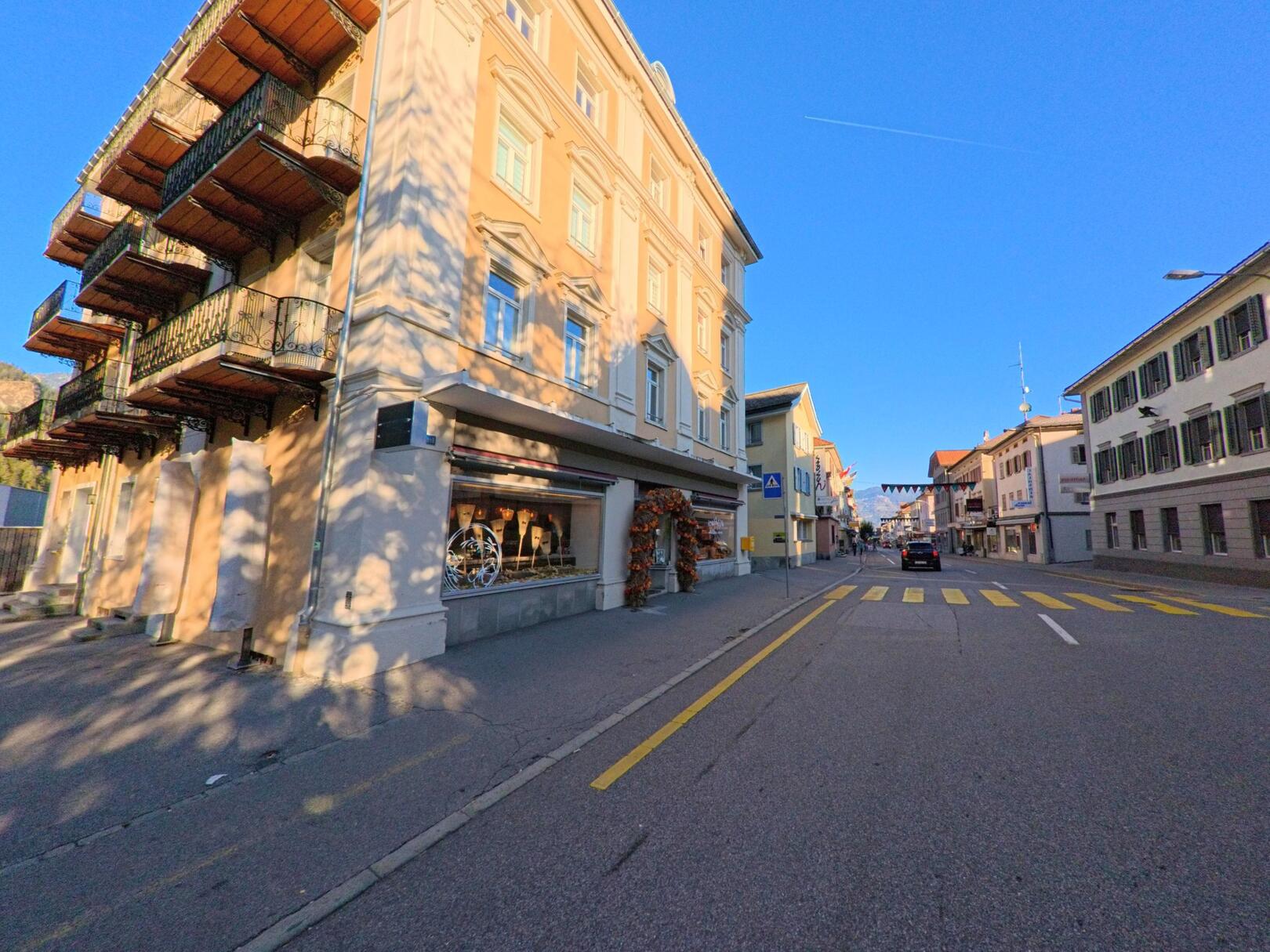 Entlang einer belebten Strasse zeigt sich in Thusis (7430, Kanton Graubünden) ein historisches Eckgebäude mit Balkonen und Ladenfronten im warmen Abendlicht.