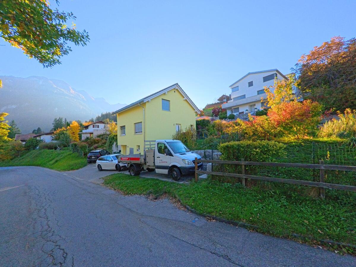 An einer ruhigen Wohnstrasse in Thusis (7430, Kanton Graubünden) steht ein gelbes Einfamilienhaus mit parkierenden Fahrzeugen, umgeben von herbstlich gefärbten Gärten und Hanglagen.