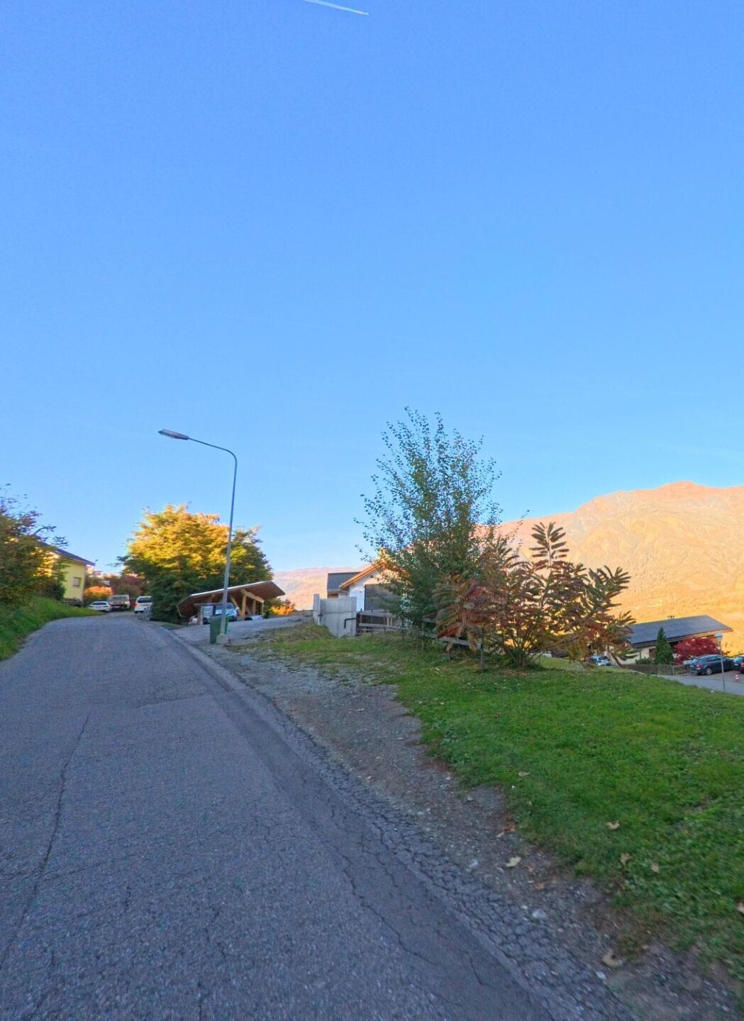 An einer leicht ansteigenden Wohnstrasse befinden sich in Thusis (7430, Kanton Graubünden) einzelne Häuser mit Gärten vor einem sonnenbeschienenen Berghang.