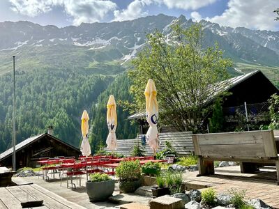 In Surses (7464, Kanton Graubuenden) bietet das Restaurant Tigia eine Terrasse mit roten Stuehlen und Sonnenschirmen vor eindrucksvoller Bergkulisse.