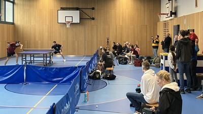 In einer Sporthalle in Affoltern am Albis (Kanton Zürich) bestreiten zwei Spieler ein Tischtennismatch, während Zuschauer und Kameras das Geschehen aufmerksam verfolgen.