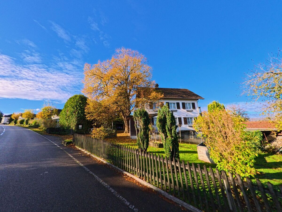 Ein freistehendes Wohnhaus mit Garten und herbstlich gefaerbten Baeumen liegt entlang einer Strasse in Tobel-Taegerschen (9555, Kanton Thurgau) bei klarem Himmel.