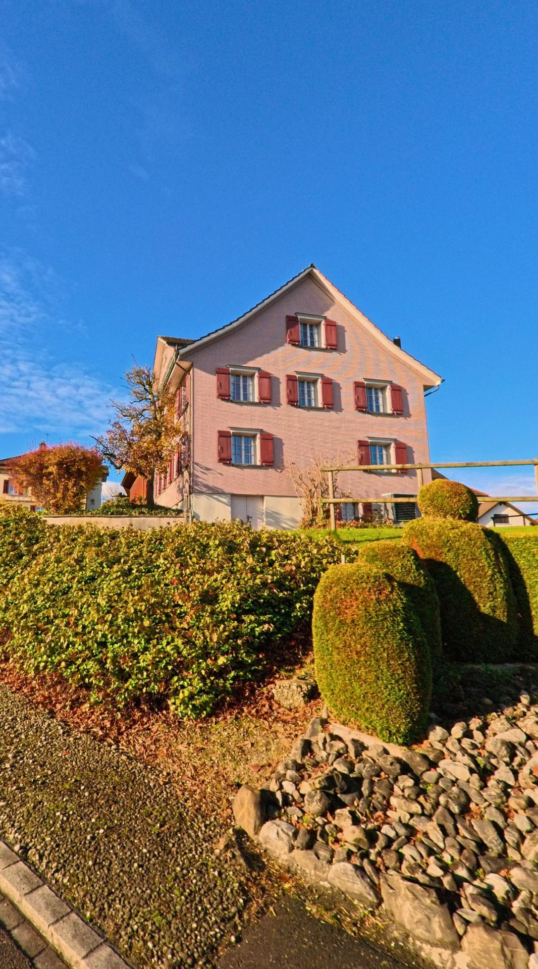Ein traditionelles Wohnhaus mit roten Fensterlaeden steht oberhalb einer gepflegten Gartenanlage in Tobel-Taegerschen (9555, Kanton Thurgau) bei klarem Himmel.