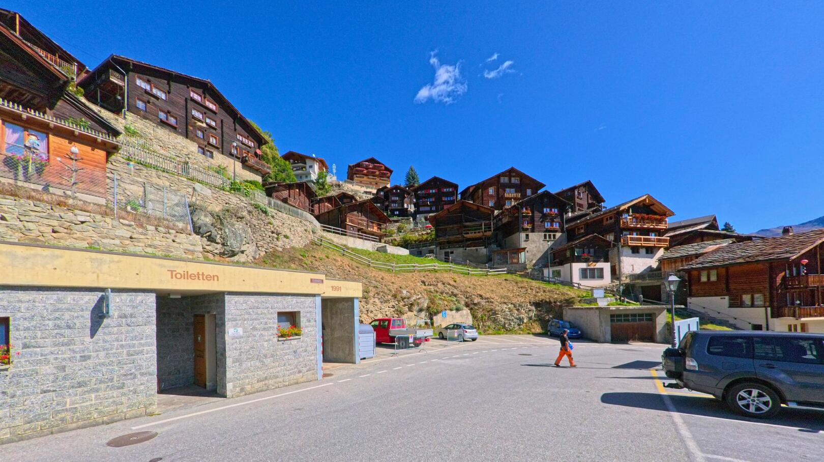 Am Hang von Törbel (3923, Kanton Wallis) reihen sich dunkle Holzhäuser eng aneinander, während im Vordergrund ein Parkplatz mit einem modernen Toilettengebäude sichtbar ist.