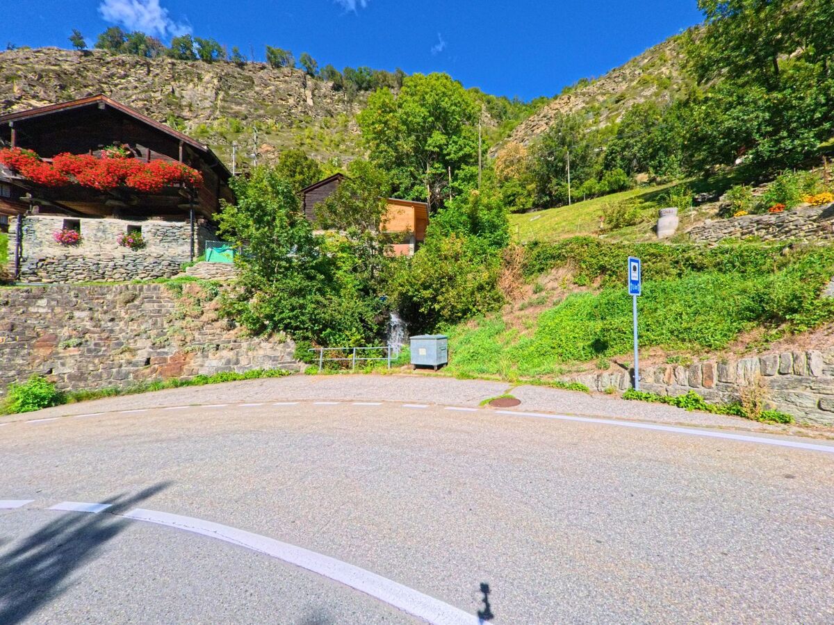 An einer Strassenkurve in Törbel (3923, Kanton Wallis) stehen Häuser mit blumengeschmückten Balkonen, daneben ein Haltstellenschild vor begrünter Hanglage.