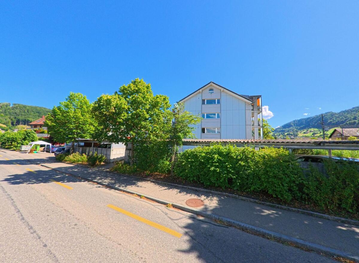 In Toffen (3125, Kanton Bern) steht ein helles Mehrfamilienhaus hinter einer begrünten Mauer entlang einer ruhigen Strasse mit Sicht auf die umliegenden Hügel.
