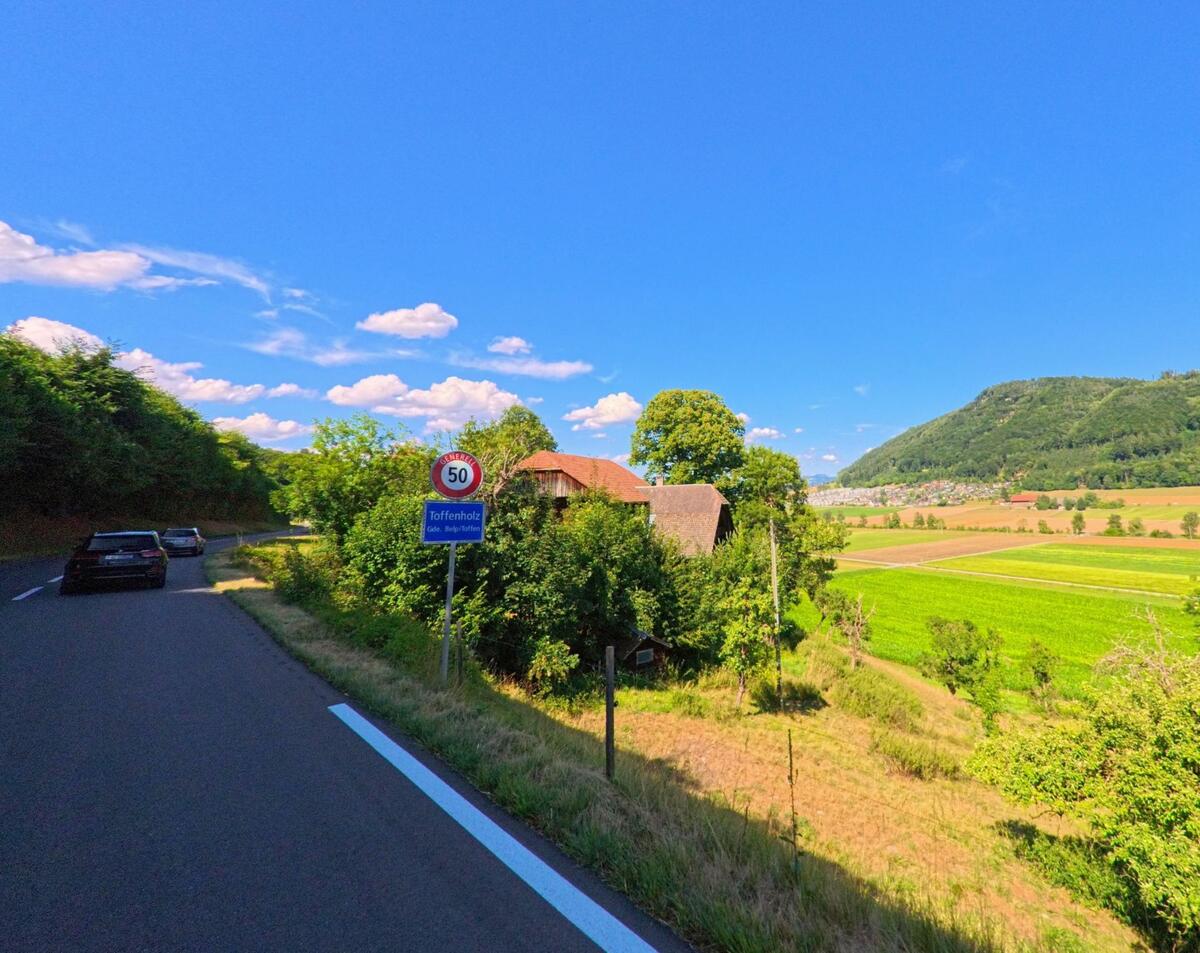 An der Ortseinfahrt von Toffenholz bei Toffen (PLZ 3125, Kanton Bern) markiert ein Verkehrsschild die Geschwindigkeitsbegrenzung, während im Hintergrund Felder und bewaldete Hügel liegen.