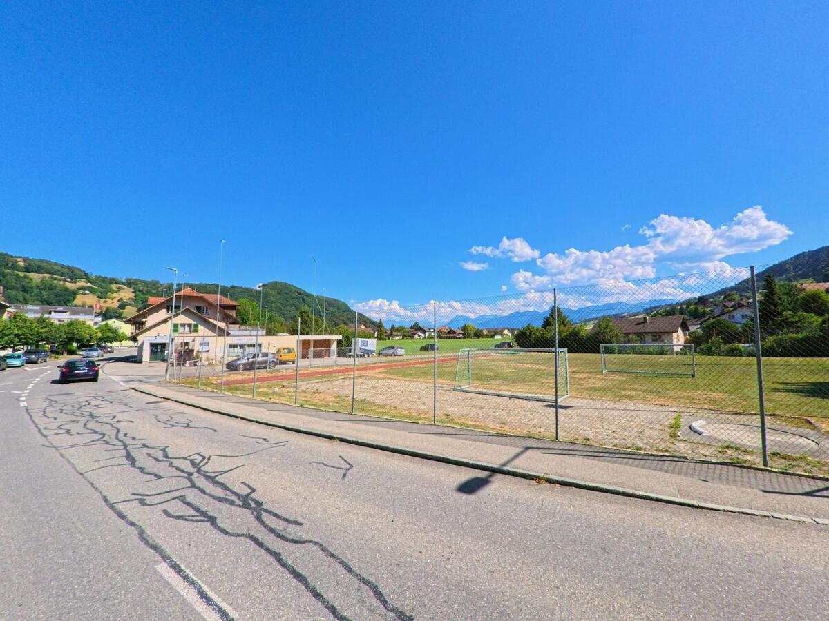 In Toffen (PLZ 3125, Kanton Bern) verläuft eine Quartierstrasse am umzäunten Fussballplatz entlang, mit Blick auf Wohnhäuser und bewaldete Hügel im Hintergrund.