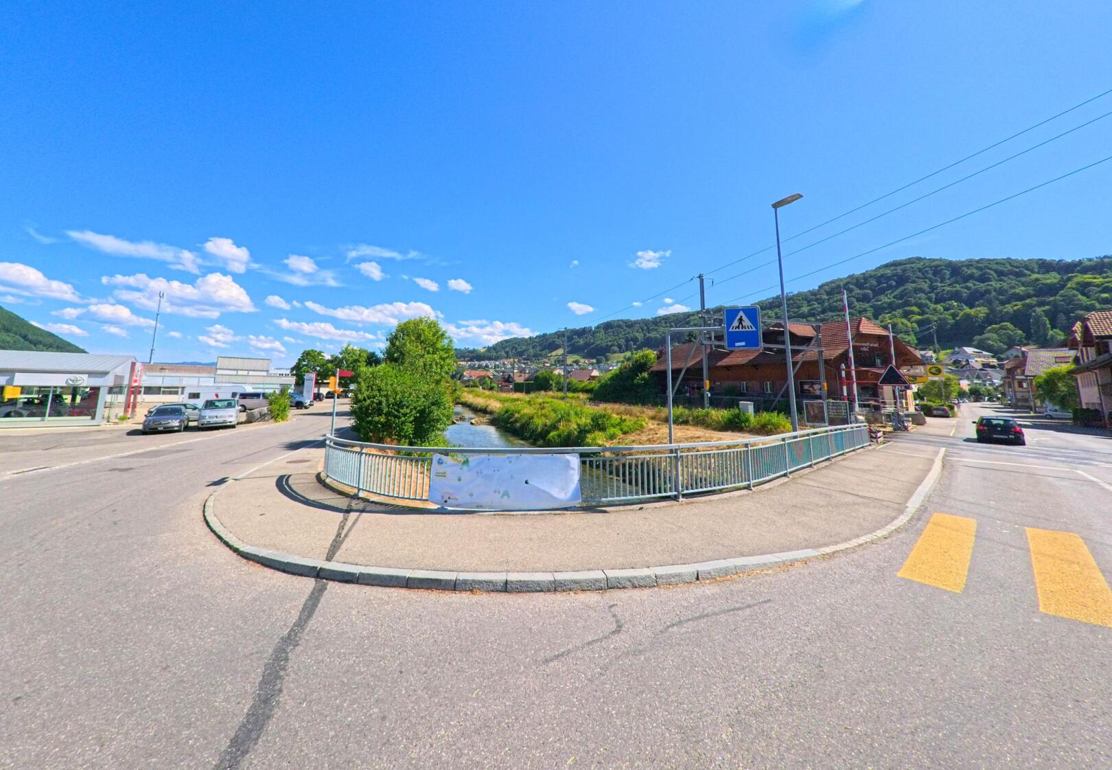 In Toffen (3125, Kanton Bern) kreuzen sich zwei Strassen an einer Brücke über einen Bach, flankiert von Wohnhäusern und einem Autohaus, mit bewaldeten Hügeln im Hintergrund.