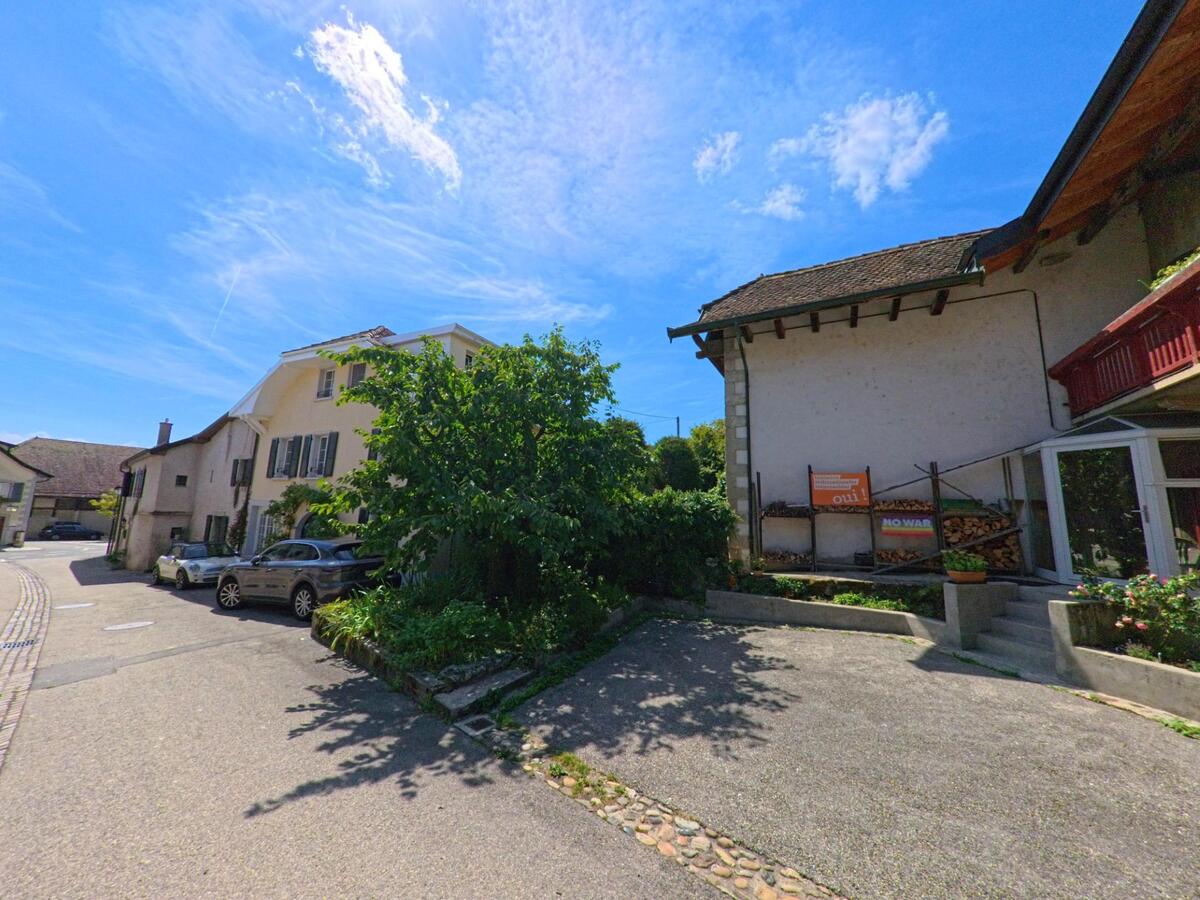 Zwischen Wohnhaeusern in Tolochenaz (1131, Kanton Waadt) fuehrt eine schmale Strasse vorbei an Parkplaetzen, Baeumen und kleinen Gartenanlagen.
