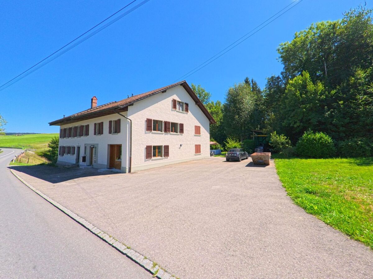 Neben einem Waldrand in Torny (1748, Kanton Freiburg) steht ein langgezogenes weisses Wohnhaus mit braunen Fensterläden und grossem Vorplatz.