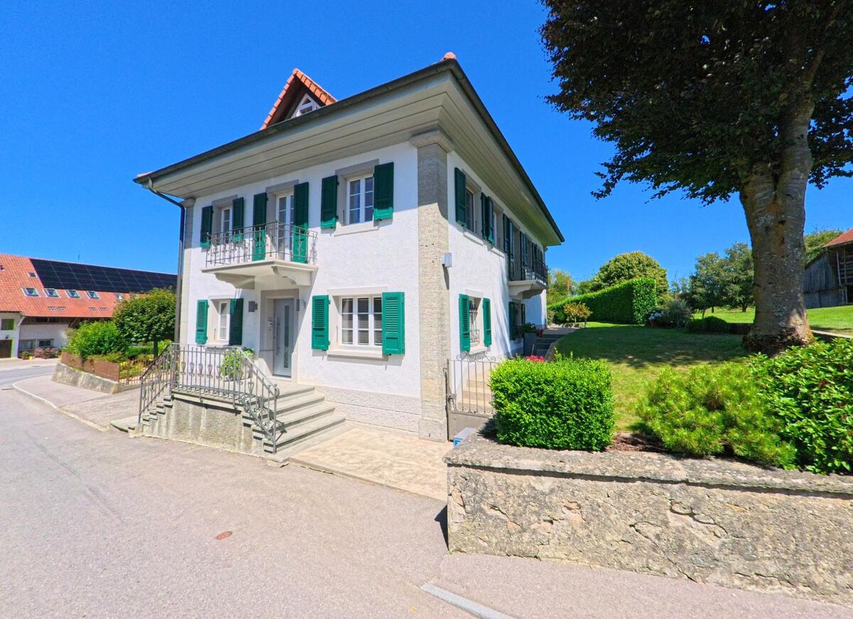 In Torny (1749, Kanton Freiburg) steht ein gepflegtes zweistöckiges Wohnhaus mit grünen Fensterläden und kleinem Balkon neben einer grün bepflanzten Gartenfläche.