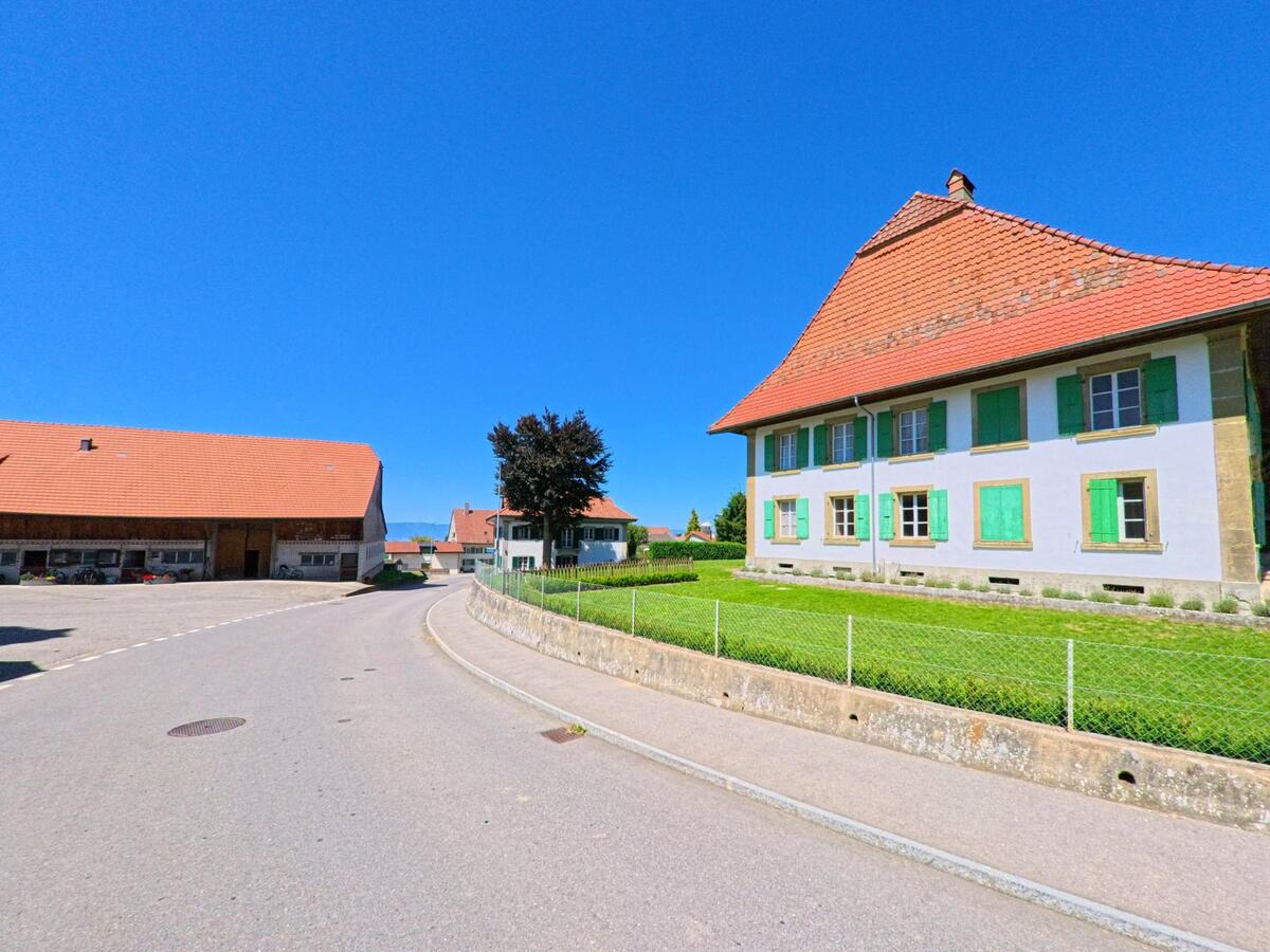 Entlang einer sanft geschwungenen Strasse in Torny (1749, Kanton Freiburg) steht ein grosses, traditionelles Bauernhaus mit grünen Fensterläden und rotem Ziegeldach.