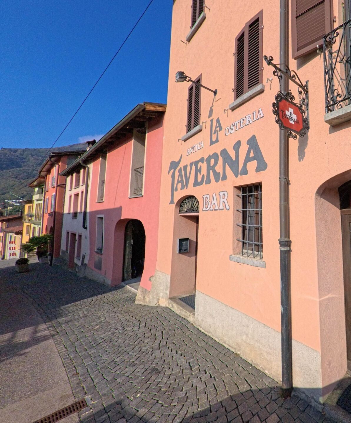 Entlang einer gepflasterten Gasse zeigt sich die Fassade einer traditionellen Taverne in Torricella-Taverne (6907, Kanton Tessin) bei sonnigem Tageslicht.