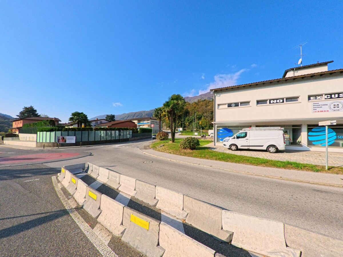 An einer verkehrsberuhigten Kreuzung mit Leitblöcken zeigt sich ein Gewerbebereich in Torricella-Taverne (6907, Kanton Tessin) vor bewaldeten Hügeln.