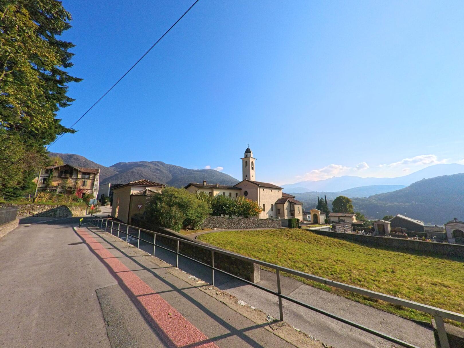 Entlang eines leicht ansteigenden Weges liegt eine Kirche mit angrenzendem Friedhof in Torricella-Taverne (6908, Kanton Tessin) vor einer hügeligen Landschaft.