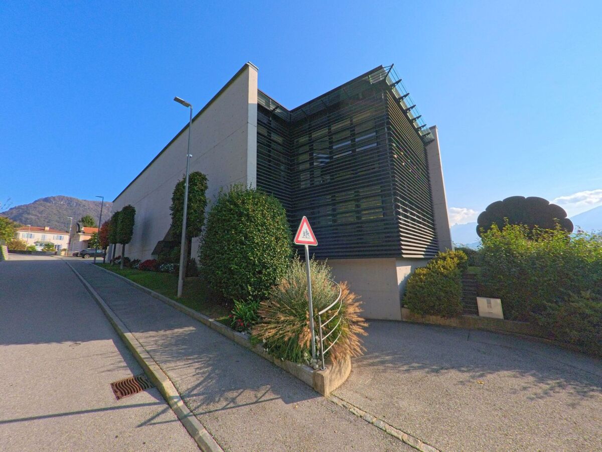 An einer leicht ansteigenden Strasse steht ein modernes Gebäude mit markanter Fassadenstruktur in Torricella-Taverne (6908, Kanton Tessin) unter klarem Himmel.