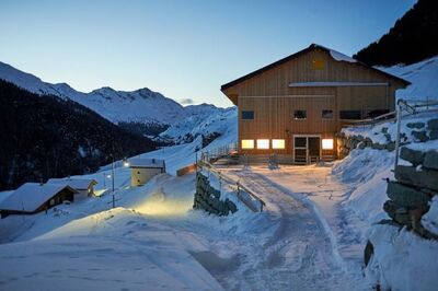 In Avers (7447, Kanton Graubuenden) leuchtet ein Holzgebaeude im Schnee, umgeben von verschneiten Bergen und einem abendlichen Himmel.
