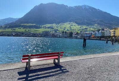 Eine rote Bank steht am Ufer eines Sees mit Blick auf Dorf und Berge in Buochs (6374, Kanton Nidwalden) und zeigt eine ruhige Landschaft.