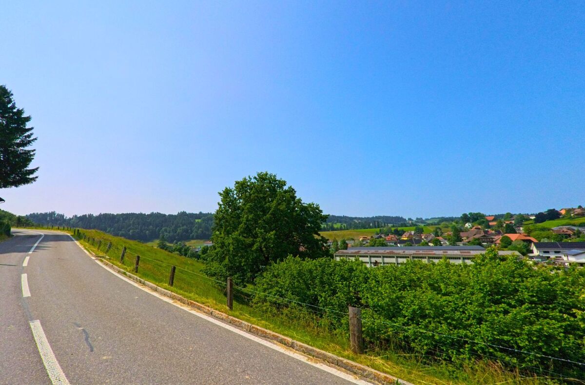 Eine sanft geschwungene Strasse führt oberhalb von Trachselwald (PLZ 3455, Kanton Bern) durch grüne Hügel mit Blick auf das Dorf und die angrenzenden Felder.