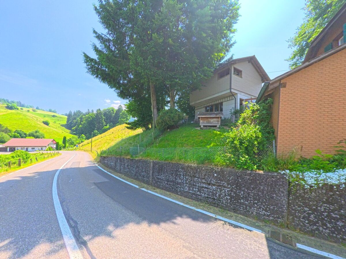Ein Wohnhaus auf erhöhter Lage mit vorgelagertem Garten steht am Waldrand von Trachselwald (3456, Kanton Bern), oberhalb einer kurvigen Landstrasse in hügeliger Landschaft.