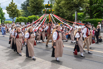 Eine Gruppe in Trachtenkleidung zieht bei einem Umzug mit Baendern und Blumenschmuck durch eine Strasse in Gurmels (1792, Kanton Freiburg).