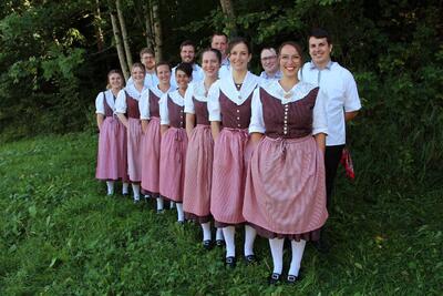 In Unteriberg (8842, Kanton Schwyz) steht die Trachtengruppe Unteriberg in traditioneller Kleidung in einer Reihe auf einer Wiese vor einem bewaldeten Hintergrund.