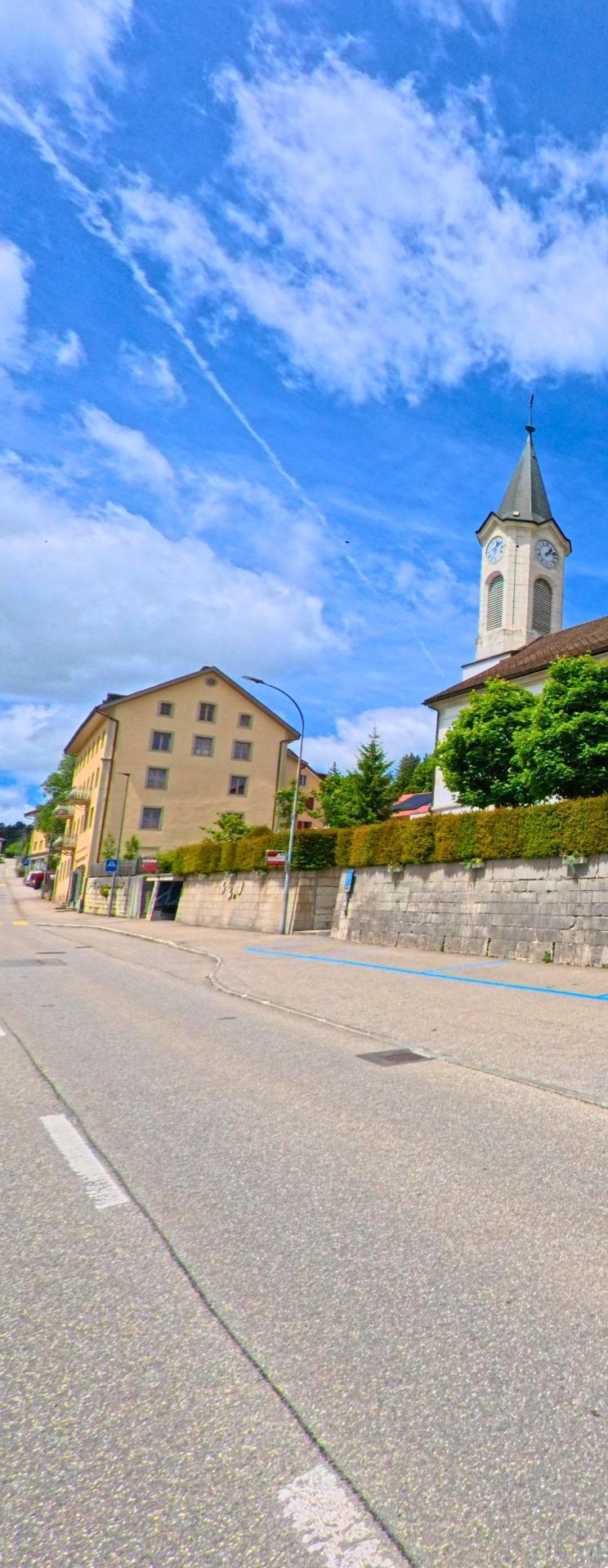 Entlang einer leicht ansteigenden Strasse prägen historische Gebäude und der Kirchturm das Ortsbild von Tramelan (2720, Kanton Bern) bei sonnigem Wetter.