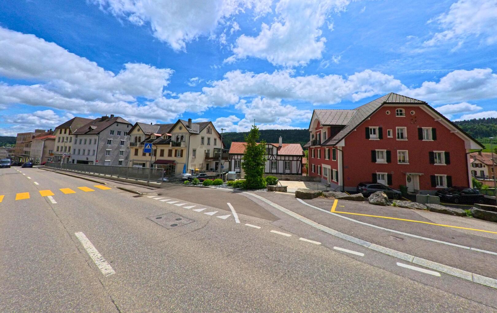 An einer übersichtlichen Kreuzung mit Fussgängerstreifen stehen Wohn- und Geschäftsgebäude im Ortszentrum von Tramelan (2720, Kanton Bern) unter einem wolkigen Himmel.