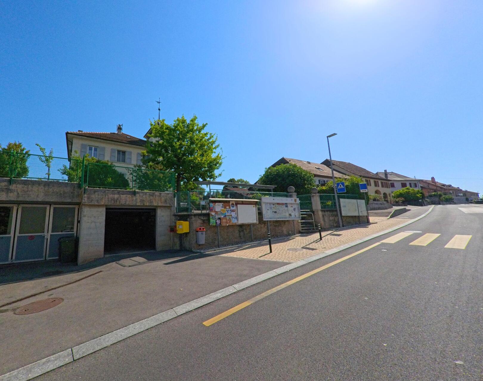 Eine Strasse mit Fussgaengerstreifen fuehrt in Treycovagnes (1436, Kanton Waadt) an Anschlagtafeln und Wohnhaeusern vorbei.