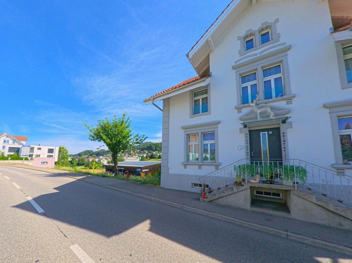 In Triengen (6234, Kanton Luzern) steht ein weiss verputztes Wohnhaus mit dekoriertem Eingangsbereich direkt an einer befahrenen Strasse.