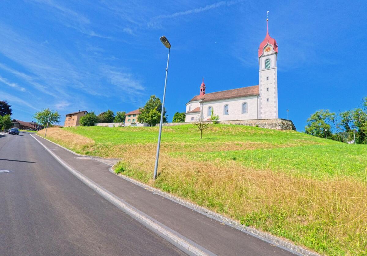 Auf einer leichten Anhöhe in Triengen (6235, Kanton Luzern) steht eine Kirche mit rotem Turmhelm, umgeben von Wiesen und angrenzenden Wohnhaeusern.