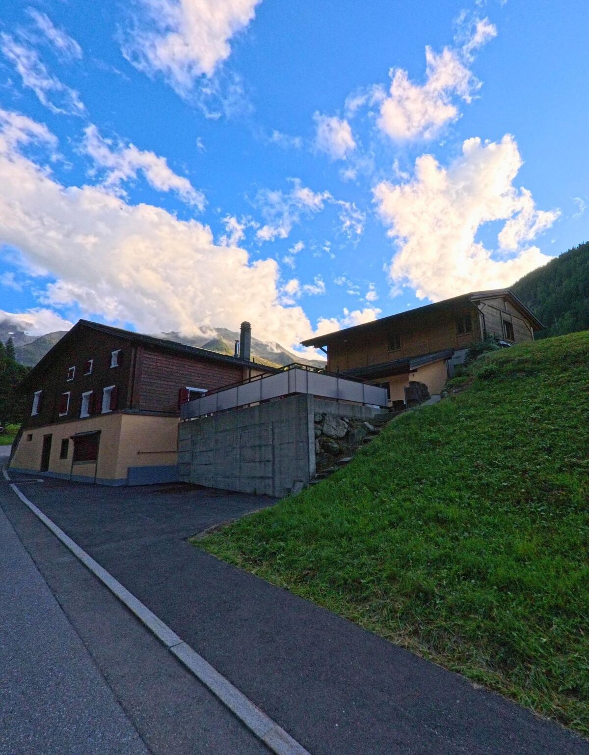 Zwei Wohnhaeuser am Hang von Trient (1929, Kanton Wallis) heben sich vor blauem Himmel mit Wolkenformationen ab.