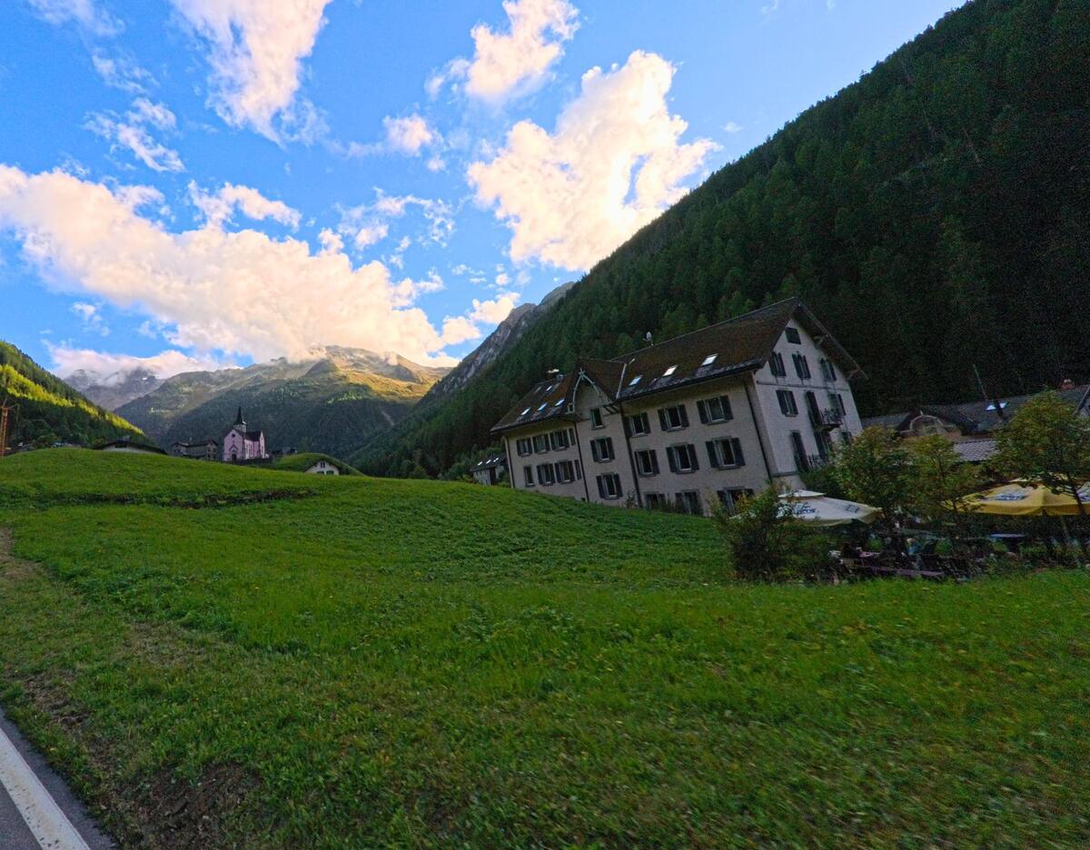 Ein grosses Wohnhaus und die Kirche von Trient (1929, Kanton Wallis) liegen am Fuss bewaldeter Haenge mit Blick auf die umliegenden Berge.
