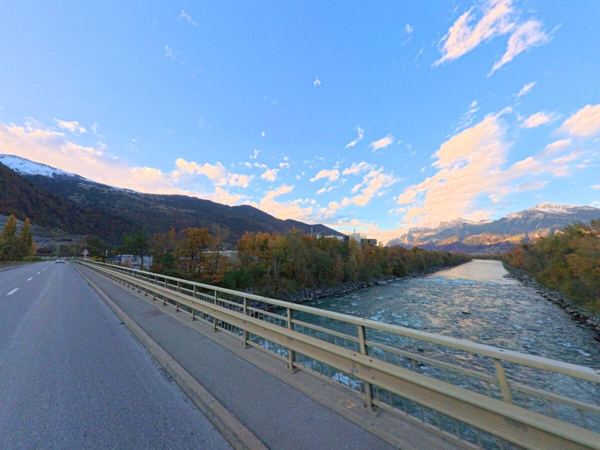 Auf einer Brücke in Trimmis (7203, Kanton Graubünden) verläuft die Strasse parallel zu einem breiten Fluss, begleitet von herbstlichen Uferbäumen und einem weiten Blick auf die umliegenden Berge.