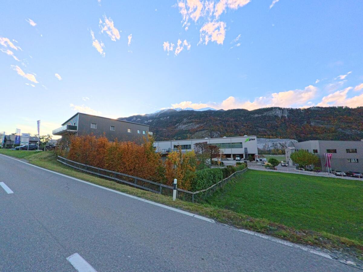 Am Rand des Gewerbegebiets von Trimmis (7203, Kanton Graubünden) verläuft eine Strasse entlang moderner Betriebsgebäude, eingerahmt von Wiesen und herbstlich gefärbten Berghängen.